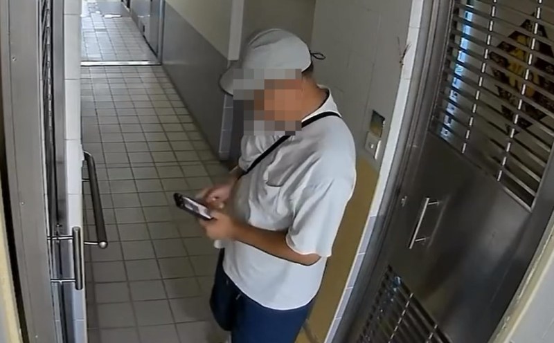 片中一名身穿白色短袖上衣、蓝色短裤、头戴浅灰色鸭舌帽的男子站在公共走廊上,他右手在裤袋位置翻找并掏出物件;其后男子疑似打开手机镜头。